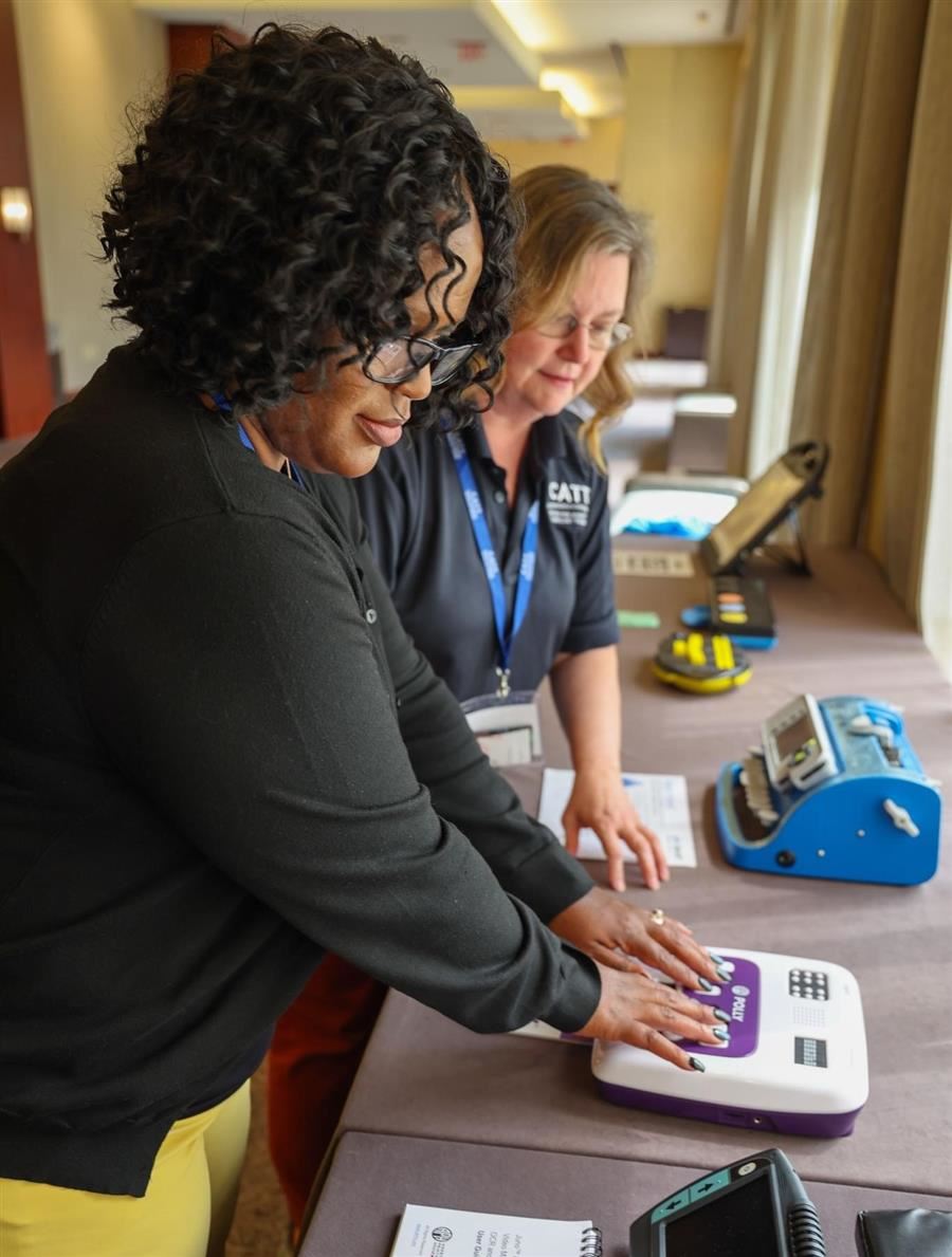 2 women use assistive technology at a table. 1 places hands on a tactile device with purple buttons.